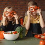 Funny boy and girl wearing apron looking through slices of pepper while preparing healthy vegetarian salad together at table with ingredients and bowls