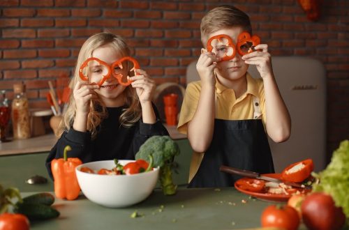 Funny boy and girl wearing apron looking through slices of pepper while preparing healthy vegetarian salad together at table with ingredients and bowls