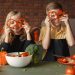 Funny boy and girl wearing apron looking through slices of pepper while preparing healthy vegetarian salad together at table with ingredients and bowls